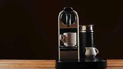 A coffee maker holds a cup, next to a frother and small pitcher, all on a black base and wood table against a dark background.