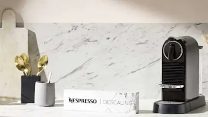 Een koffiezetapparaat met een witte mok eronder, naast een melkopschuimer en een klein melkkannetje op een houten tafel.
