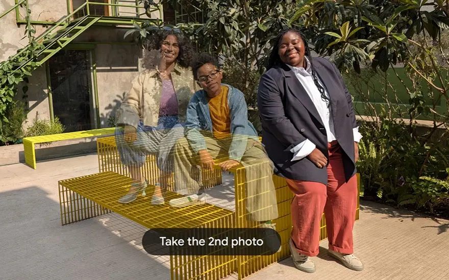 Drie vrouwen poseren vrolijk op gele draadmeubilair in een zonnige buitenruimte. Moderne tuinset met geometrisch design en groene planten.