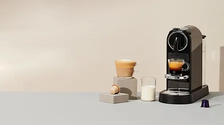 Zwarte koffiemachine met kopje espresso, melkkan en houten bak op grijze blokken tegen een beige achtergrond.
