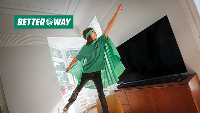 A person in a green cape and mask stands with arm raised in a room with a TV. A logo is in the upper left corner.