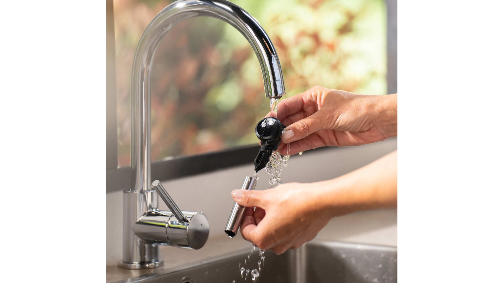 Een persoon spoelt een zwart apparaat en een zilveren buis af onder een glanzende, metalen kraan in een gootsteen.