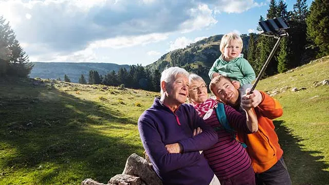 Een familie maakt een selfie met een selfiestick in een bergachtig gebied met gras en bomen.