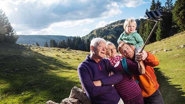 A family takes a selfie outdoors. A child sits on a man's shoulders, with mountains and trees in the background.