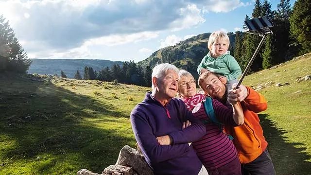 A family takes a selfie outdoors. A child sits on a man's shoulders, with mountains and trees in the background.
