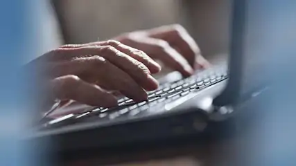 Twee handen typen op een zwart laptoptoetsenbord. De laptop staat op een lichte ondergrond.