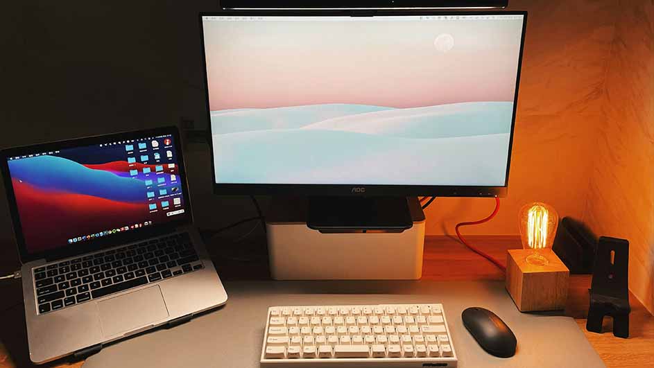 Een bureau met een laptop, monitor, toetsenbord, muis, lamp en telefoonstandaard. De monitor toont een roze-witte afbeelding.