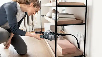 Een vrouw knielt en stofzuigt een zwarte metalen stellingkast met houten planken, waarop boeken en dozen staan.