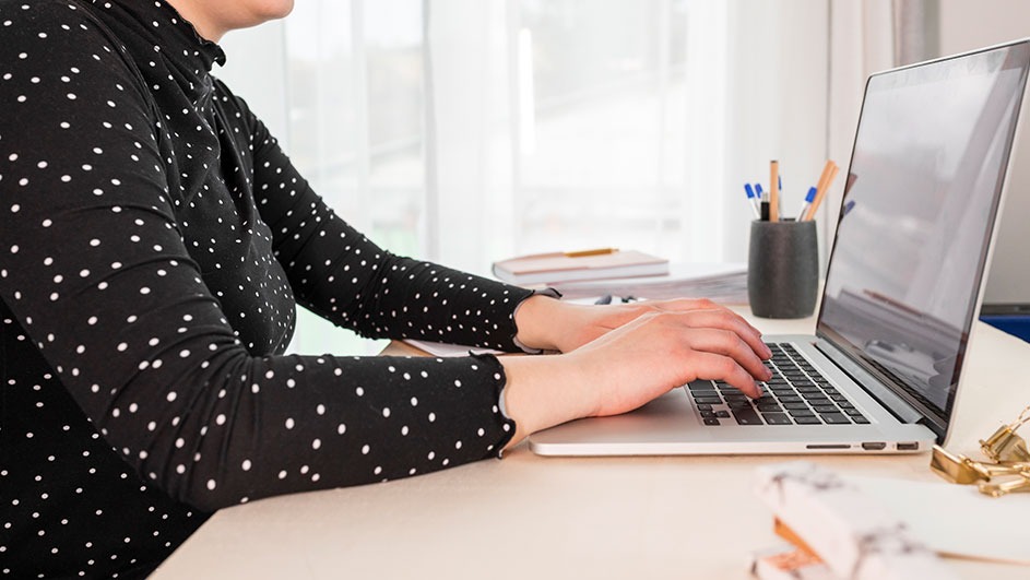 Person types on a silver laptop at a desk. A black polka dot shirt is visible. Pens and notebooks are in the background.