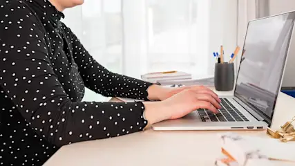 Person types on a silver laptop at a desk. A black polka dot shirt is visible. Pens and notebooks are in the background.
