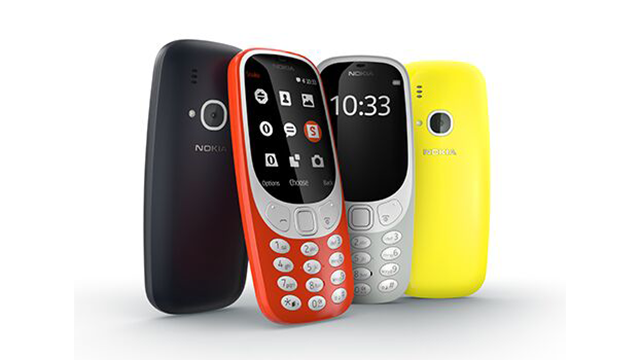 Vier Nokia 3310-telefoons in zwart, oranje, grijs en geel staan rechtop op een witte achtergrond.