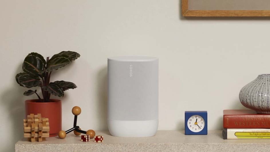 Planten, een speaker, een blauwe klok, boeken en een vaas staan op een plank tegen een witte muur.