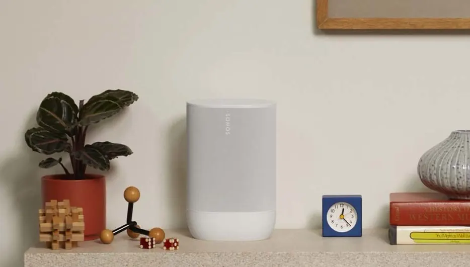 Planten, een speaker, een blauwe klok, boeken en een vaas staan op een plank tegen een witte muur.