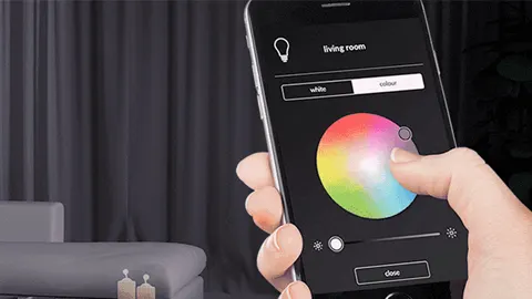 Een hand bedient een smartphone-app met een kleurenpalet voor verlichting, in een kamer met een grijze bank en kaarsen.