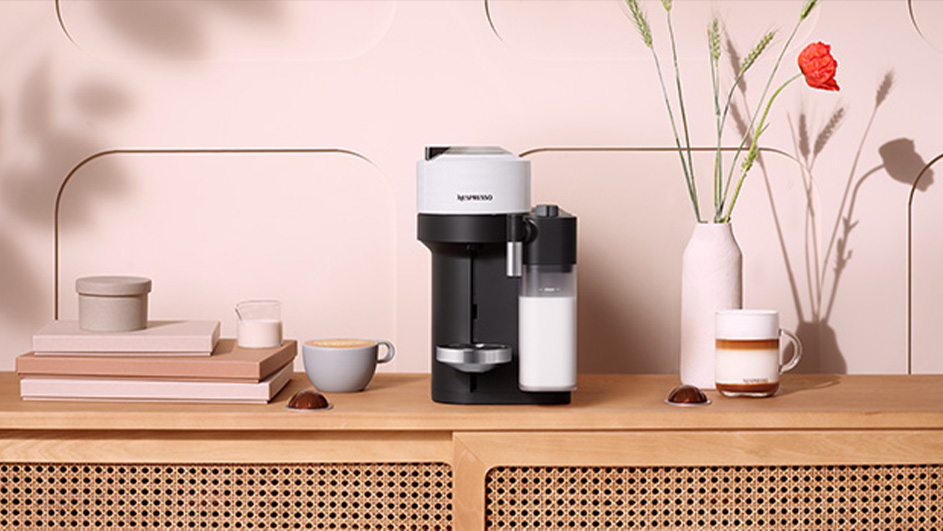 Koffiezetapparaat naast kopjes, melkkan, bloemen in vaas op houten kast tegen een roze achtergrond.