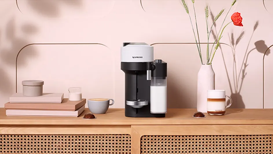 Koffiezetapparaat naast kopjes, melkkan, bloemen in vaas op houten kast tegen een roze achtergrond.