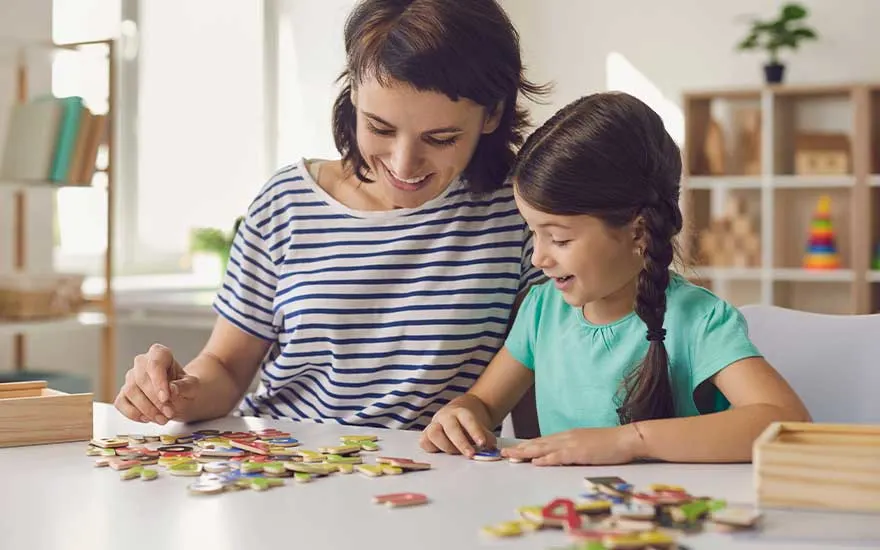 Een moeder en dochter zitten samen aan tafel en spelen lachend met kleurrijke magnetische cijfers en letters in een lichte woonkamer.