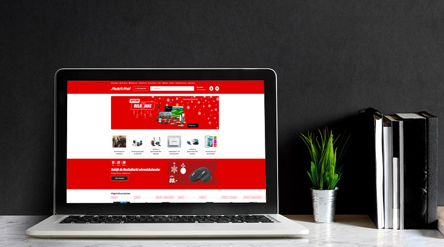 Laptop met rode webpagina op een tafel, naast een plant in een pot en boeken tegen een zwarte achtergrond.