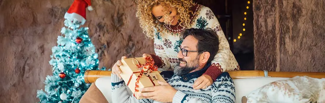 Een vrouw geeft glimlachend een ingepakt kerstcadeau aan een man op de bank, naast een kleine verlichte kerstboom in een knusse woonkamer.
