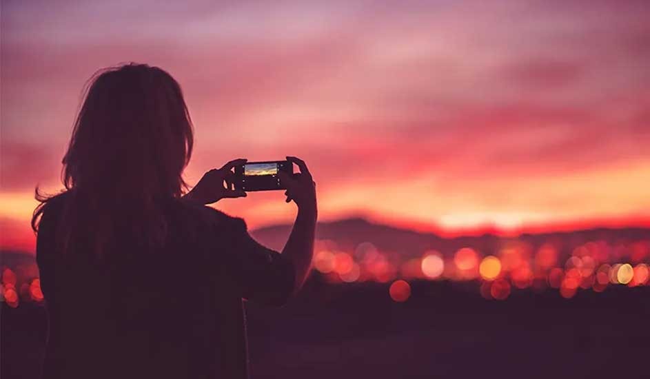 A person photographs a colorful sunset with a smartphone. The brand of the phone is not visible in the picture.