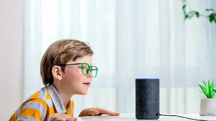 Een kind met een bril kijkt naar een zwarte slimme luidspreker op een witte tafel, met een plant op de achtergrond.