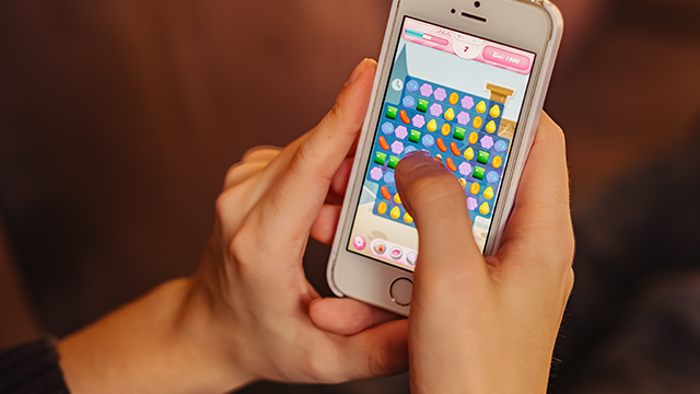 Een telefoon wordt vastgehouden met een spelletje met gekleurde snoepjes zichtbaar op het scherm tegen een bruine achtergrond.