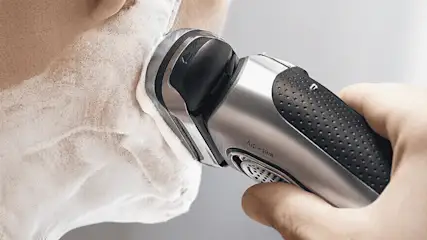 Een zilverkleurig elektrisch scheerapparaat, vastgehouden door een hand, wordt gebruikt op een gezicht bedekt met wit scheerschuim.