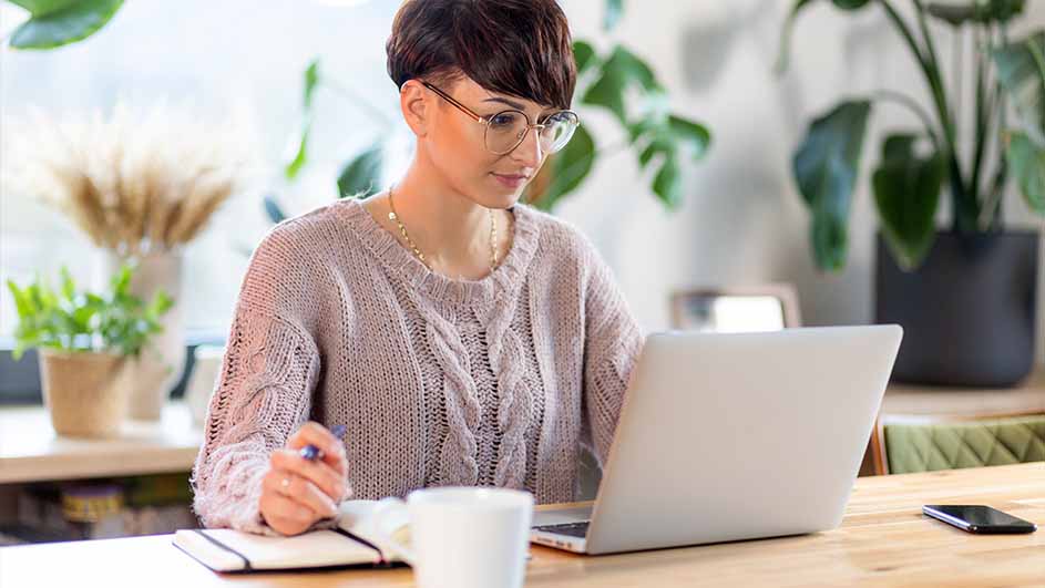 Vrouw met bril werkt aan een laptop met een notitieboek en een pen op een houten tafel, met planten op de achtergrond.