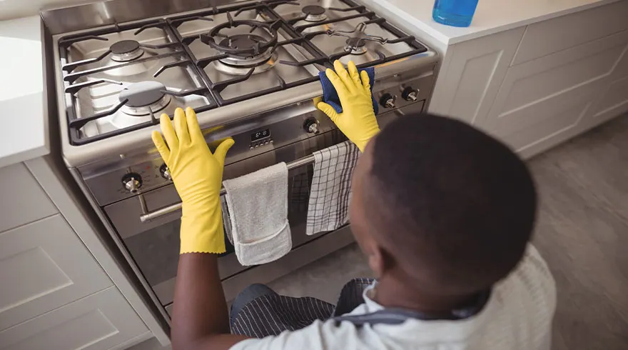 Een persoon met gele handschoenen maakt een roestvrijstalen fornuis schoon met een blauwe doek in een keuken.
