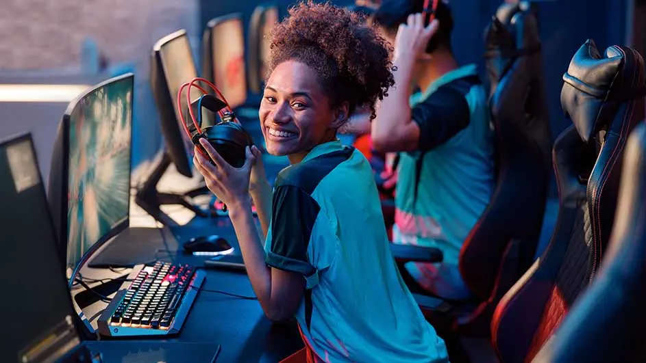 Een glimlachende persoon houdt een headset vast in een gamingkamer met computers en stoelen.