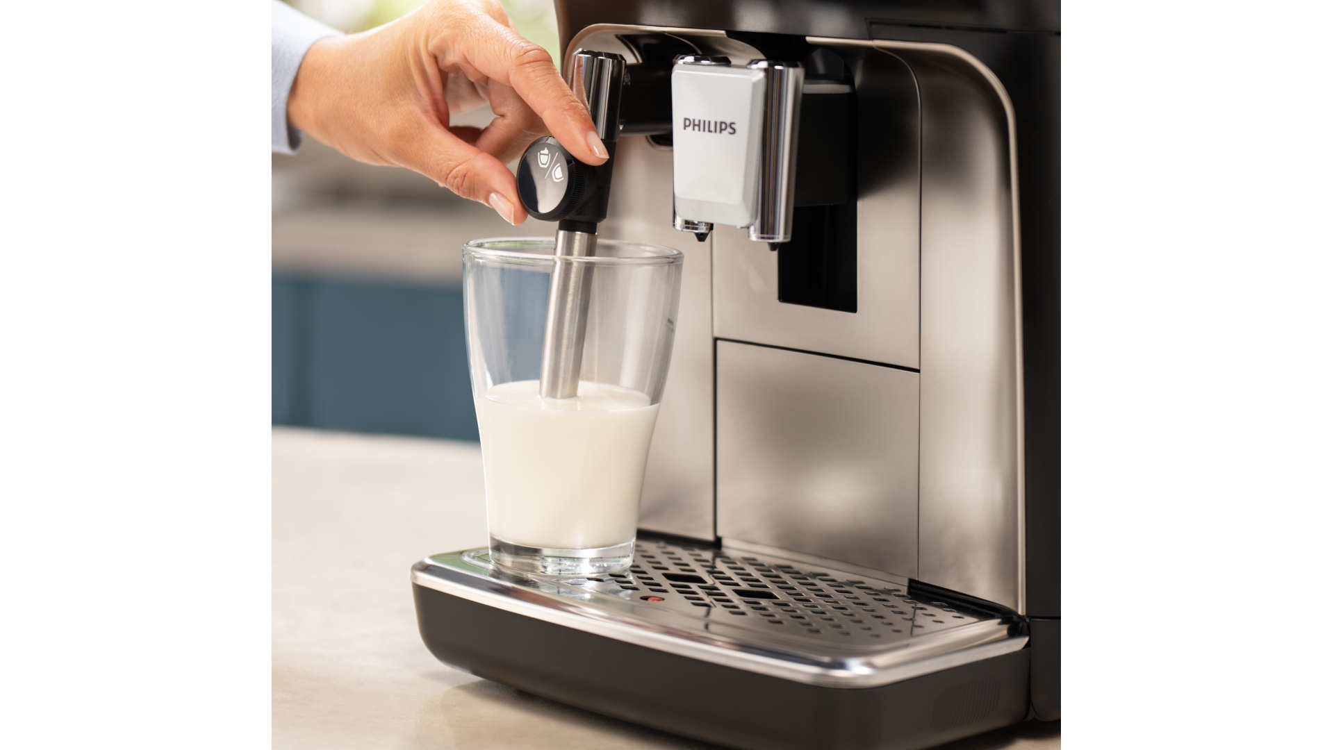 Een hand bedient een Philips-melkopschuimer boven een glas melk, naast een koffiezetapparaat.
