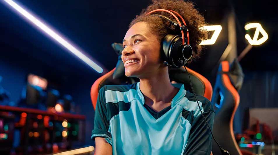 Een glimlachende persoon met een blauw-zwart shirt en een oranje koptelefoon zit in een gamestoel. Neonlichten op de achtergrond.