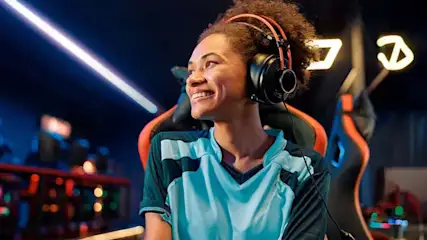 Een glimlachende persoon met een blauw-zwart shirt en een oranje koptelefoon zit in een gamestoel. Neonlichten op de achtergrond.