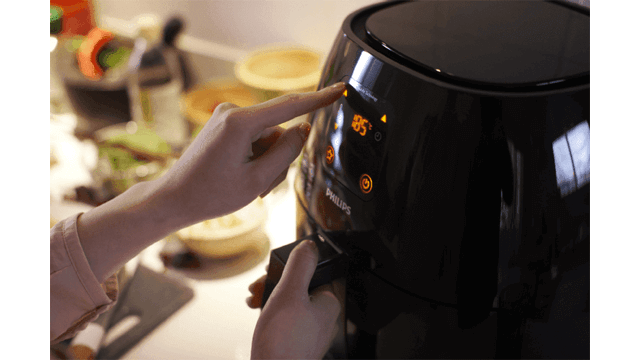 Een persoon bedient een zwarte Philips airfryer met een digitaal display dat 85 graden Celsius weergeeft.