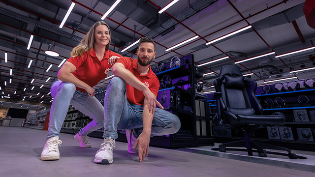 Twee mensen in rode shirts en jeans zitten gehurkt in een winkel met rekken vol elektronica en een zwarte bureaustoel.