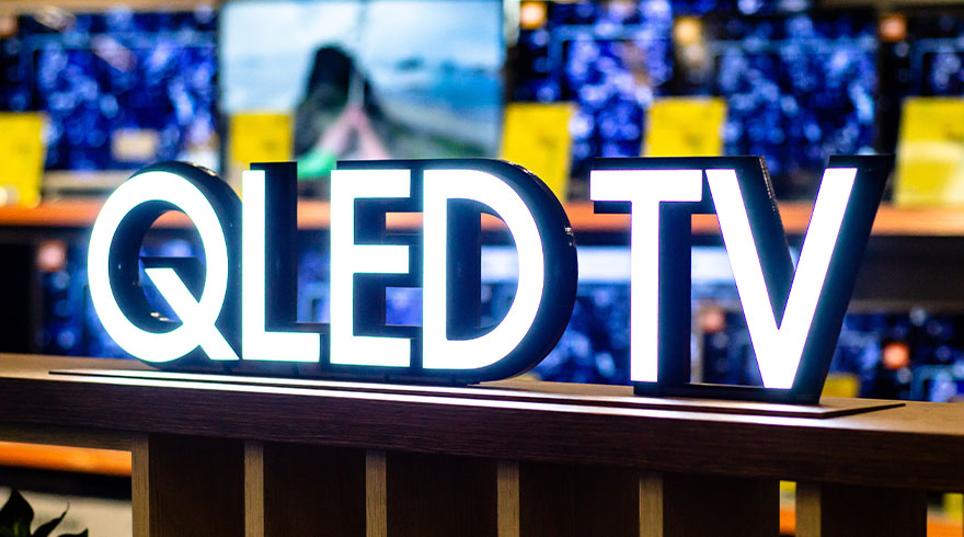Grote, witte letters "QLED TV" op een bruine plank. Achtergrond toont schermen met blauwe en gele kleuren.