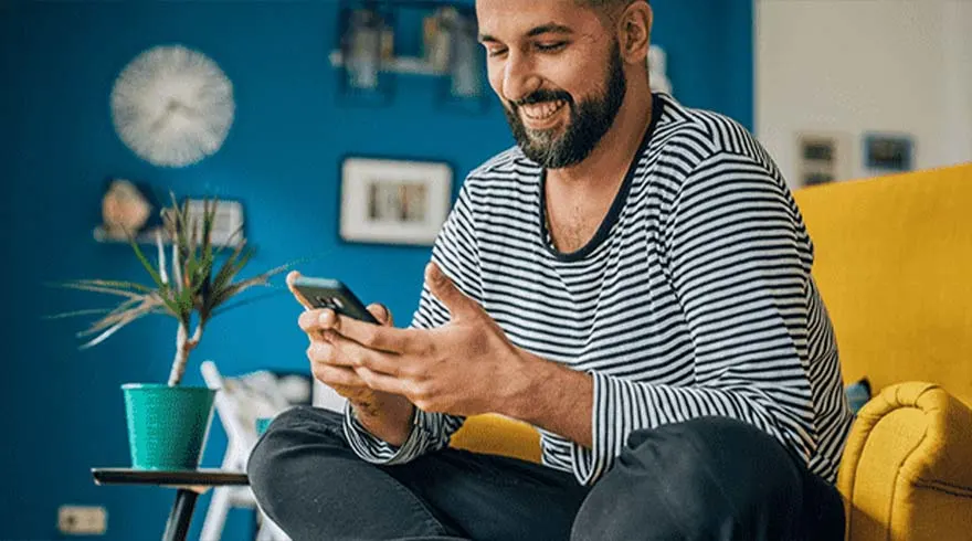 Een man zit op een gele stoel met een telefoon in zijn handen, gekleed in een gestreept shirt en een blauwe achtergrond.