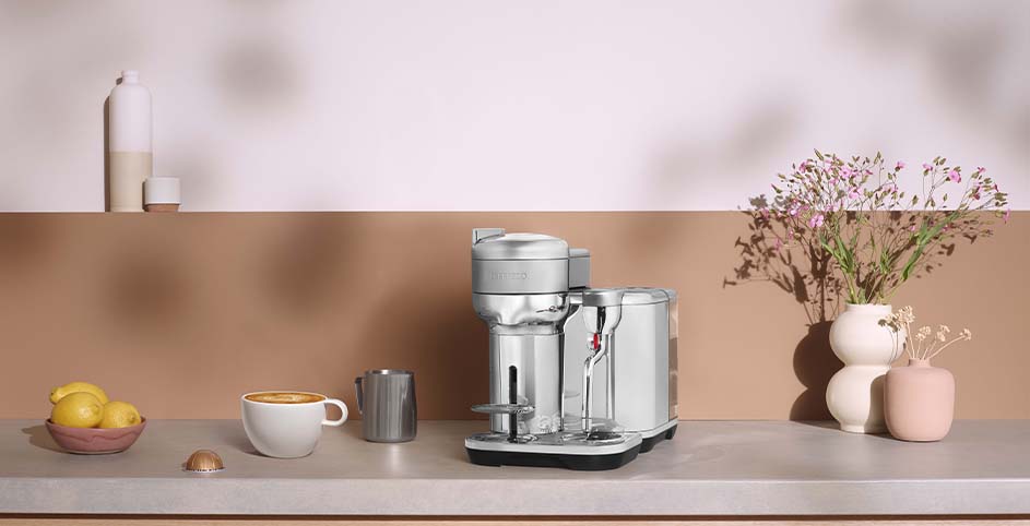 Een zilverkleurig koffiezetapparaat op een aanrecht met een kop koffie, citroenen, een melkkan en bloemen.