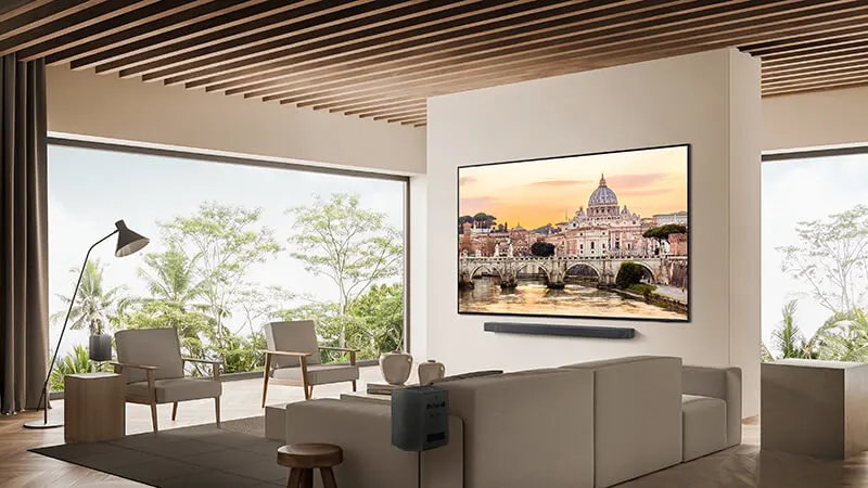 Woonkamer met beige bank, stoelen, lamp en groot scherm met stadsgezicht. Buiten zicht op bomen door ramen.