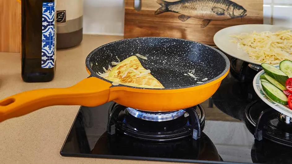 Een oranje koekenpan op een zwart gasfornuis met een omelet, naast borden met pasta en komkommer.