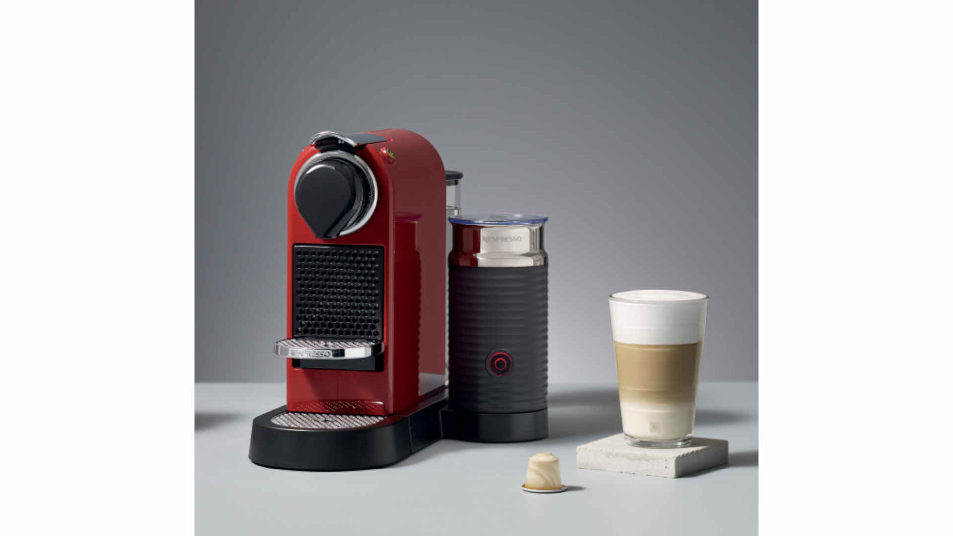 Een rode Nespresso-machine naast een melkopschuimer en een glas latte macchiato op een grijze achtergrond.