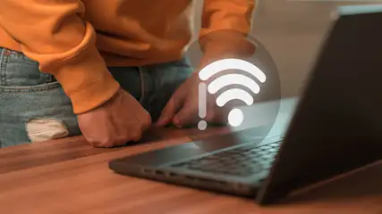 Een persoon in een oranje trui en gescheurde jeans staat bij een laptop met een wifi-symbool op een houten tafel.
