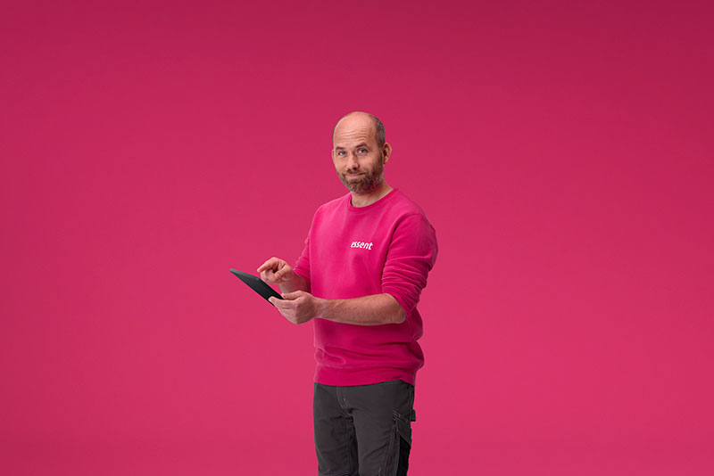Een man met een roze trui en grijze broek gebruikt een zwarte tablet tegen een roze achtergrond.