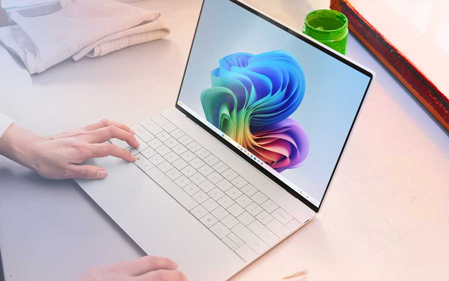 Een witte laptop met bijna randloos scherm staat op een bureau, iemand rust de handen op het toetsenbord naast opgevouwen textiel en verfbenodigdheden.