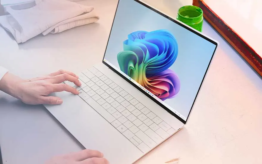 Een witte laptop met bijna randloos scherm staat op een bureau, iemand rust de handen op het toetsenbord naast opgevouwen textiel en verfbenodigdheden.