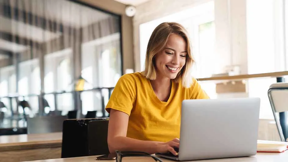 Een vrouw in een geel shirt zit lachend achter een laptop op een licht bureau. Achter haar is een kantoorruimte met ramen.