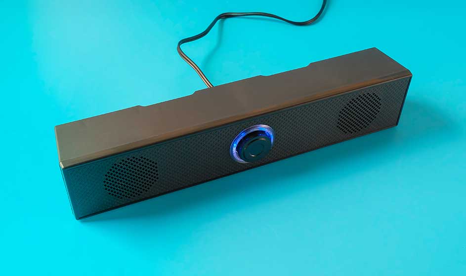 Een compacte zwarte speakerbalk ligt op een felblauwe ondergrond; in het midden gloeit een ronde knop met blauwe ring.