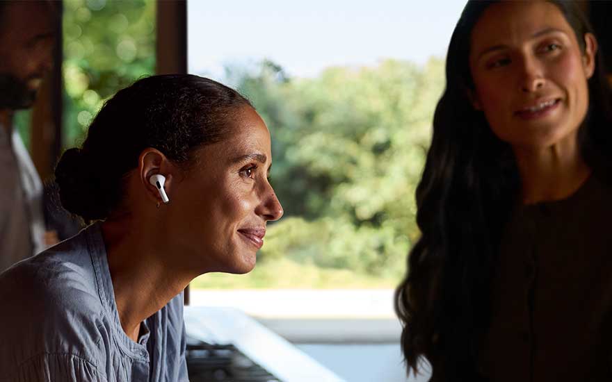 Indoors, a smiling person wears white wireless earbuds, with another person and a bright green outdoor view softly blurred in the background.