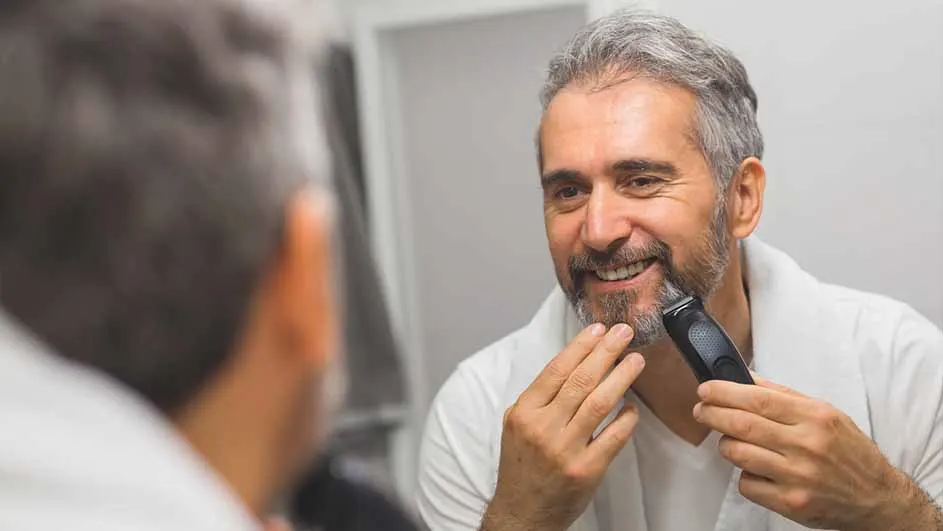 Een man scheert zijn baard met een zwarte trimmer voor een spiegel in een lichte kamer.