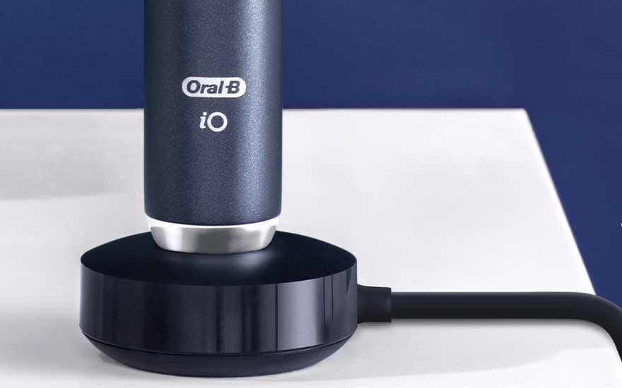 An Oral-B iO toothbrush stands upright on a black charging base with cable, placed on a white surface before a blue wall.